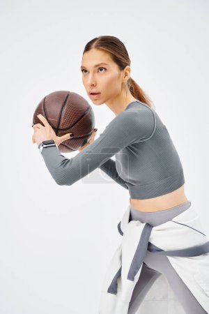 Una joven deportista en ropa activa sosteniendo una pelota de baloncesto en sus manos sobre un fondo gris.