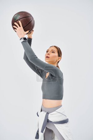 A sporty young woman gracefully holds a basketball in her right hand against a grey background.