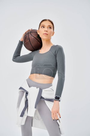 A sporty young woman confidently holds a basketball in her right hand, showcasing her athletic prowess on a grey background.
