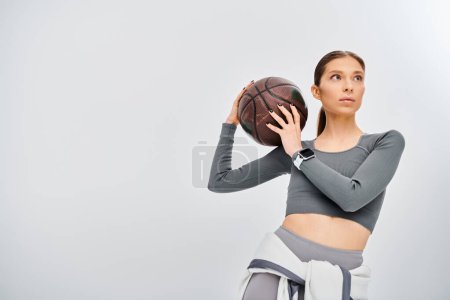 Photo for A sporty young woman holds a basketball in her right hand, showcasing grace and strength on a grey background. - Royalty Free Image