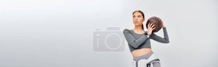 Photo for A sporty young woman elegantly holds a basketball in her right hand against a grey background. - Royalty Free Image