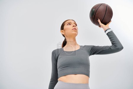 Eine sportliche junge Frau in aktiver Kleidung hält vor grauem Hintergrund einen Basketball in die Höhe.