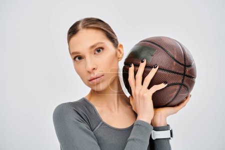 A sporty young woman in active wear holding a basketball over her face on a grey background.