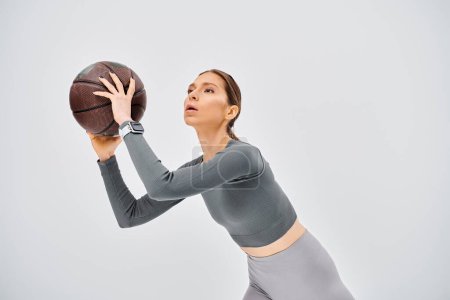 A sporty young woman gracefully holds a basketball in her right hand against a neutral grey background.