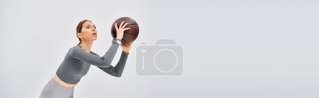 A sporty young woman in active wear confidently holds a basketball in her right hand against a grey background.