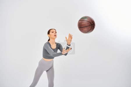 Aktive junge Frau in grauem Top dribbelt gekonnt Basketball in spielerischer Manier auf grauem Hintergrund.