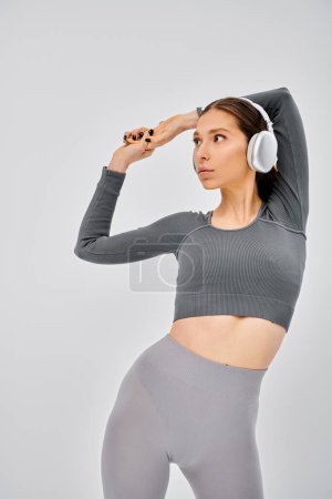 Eine sportliche junge Frau in aktiver Kleidung posiert, während sie über Kopfhörer auf grauem Hintergrund Musik hört..