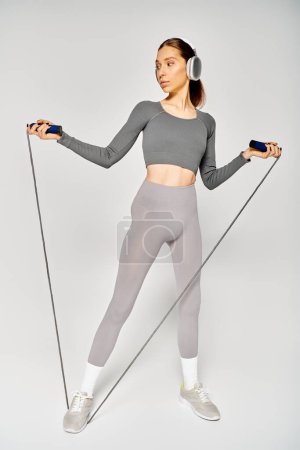 Sportliche junge Frau in aktiver Kleidung, hält Springseil in der Hand, hört Musik auf grauem Hintergrund.