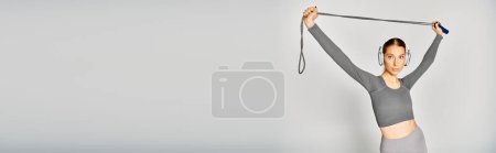 A sporty young woman in grey shirt confidently holds a skipping rope above her head against a grey background.