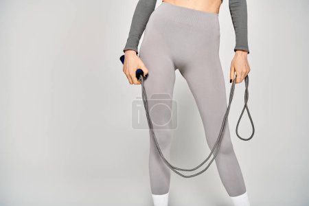 Eine sportliche junge Frau in grauer Hose steht selbstbewusst mit einem Springseil vor grauem Hintergrund..