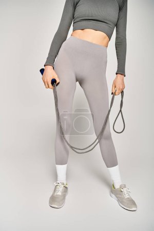 Une jeune femme sportive en tenue active tenant une corde à sauter dans ses mains sur un fond gris.
