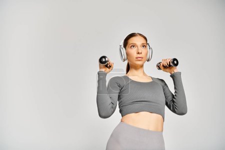 Eine sportliche junge Frau in aktiver Kleidung hält zwei Kurzhanteln in der Hand und trägt Kopfhörer auf grauem Hintergrund.