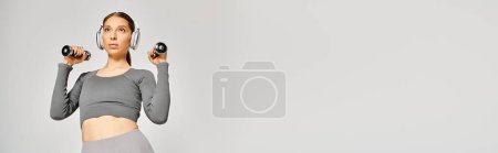 Téléchargez les photos : A sporty young woman in active wear is exercising with dumbbells against a grey background. - en image libre de droit