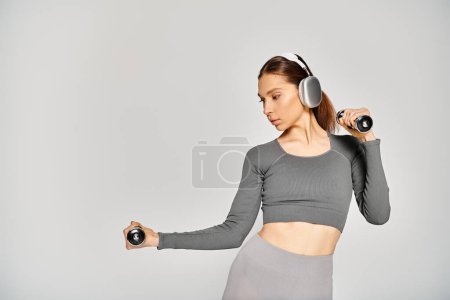 Foto de Una joven deportista sostiene pesas mientras usa auriculares, exudando concentración y determinación sobre un fondo gris. - Imagen libre de derechos