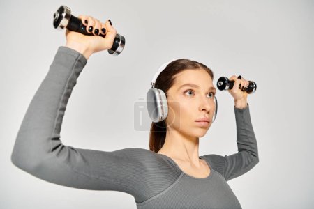 A sporty young woman wearing headphones, holding two dumbbells, exuding strength and determination on a grey background.