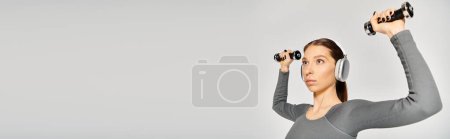 Photo for A sporty young woman holds two dumbbells in front of her head, showcasing her strength and balance. - Royalty Free Image