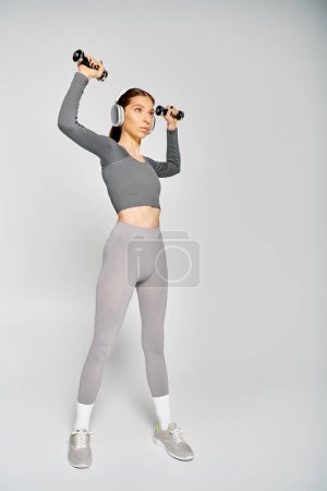 Eine sportliche junge Frau in aktiver Kleidung trainiert mit Kurzhanteln vor grauem Hintergrund.