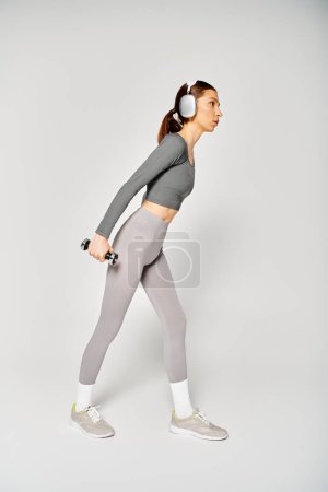 Téléchargez les photos : Une jeune femme sportive en tenue active avec des écouteurs travaillant énergiquement sur un fond gris. - en image libre de droit