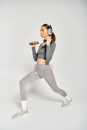A sporty young woman in active wear is energetically doing exercises while wearing headphones on a grey background.