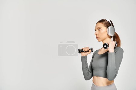 A sporty young woman holding a pair of dumbbells in active wear, showcasing strength and fitness on a grey background.