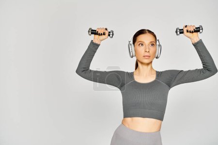Sportliche junge Frau in aktiver Kleidung hört Musik mit Kopfhörern, während sie zwei Kurzhanteln auf grauem Hintergrund hält.