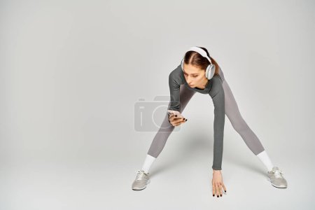 Téléchargez les photos : Une jeune femme sportive en tenue grise et blanche se concentre sur son écran de téléphone portable sur un fond gris. - en image libre de droit