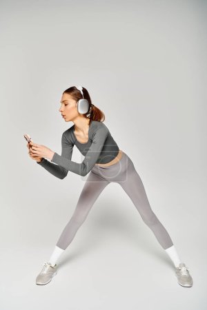 Eine sportliche junge Frau in grauer Aktivkleidung strahlt Zuversicht und Anmut aus, als sie sich auf einem neutralen grauen Hintergrund bewegt.