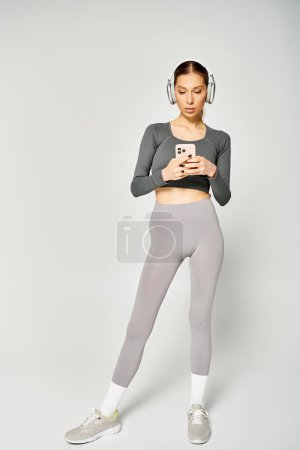 Eine sportliche junge Frau in aktiver Kleidung mit Kopfhörern blickt vor grauem Hintergrund auf ihr Handy..