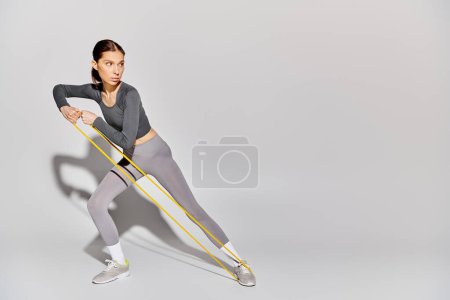 Photo for A sporty young woman in active wear confidently working out with resistance band on a grey background. - Royalty Free Image