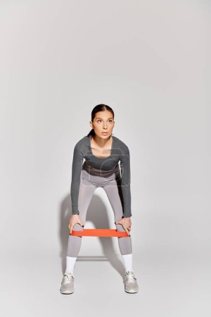 Téléchargez les photos : Une jeune femme sportive s'entraîne avec une bande de résistance sur fond gris. - en image libre de droit