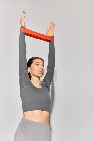 A sporty young woman in a gray top exercising out with resistance band against a light grey backdrop.