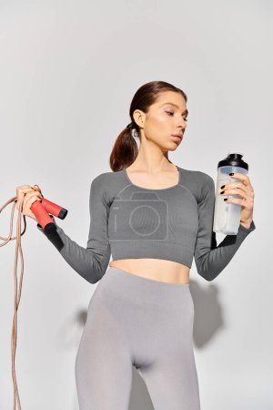 A sporty young woman in active wear holds a water bottle and jump rope, ready for a workout on a grey background.