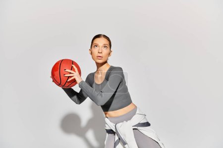 A sporty young woman in active wear holding a basketball in her right hand against a grey background.