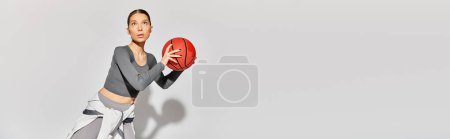 A sporty young woman in active wear holding a red basketball in her right hand against a grey background.