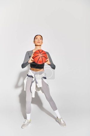 A sporty young woman elegantly holds a basketball in her right hand against a grey background.