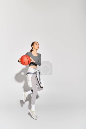 A sporty young woman in active wear gracefully holds a basketball against a grey background.