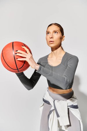 Una joven deportista en ropa activa sostiene con elegancia una pelota de baloncesto en su mano derecha sobre un fondo gris.