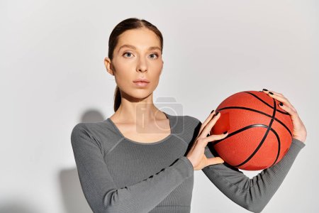 Photo for A sporty young woman in active wear gracefully holds a basketball in her hands on a grey background. - Royalty Free Image