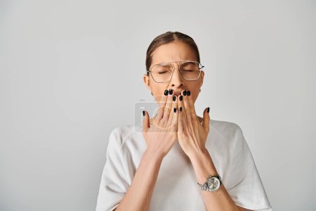 A young woman in a white t-shirt and glasses covering her mouth with her hands on a grey background.
