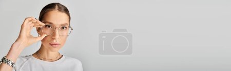 Téléchargez les photos : Une jeune femme en chemise blanche et lunettes pose sur un fond gris, respirant un sentiment de sophistication et d'intelligence. - en image libre de droit