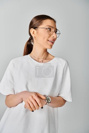 Una joven con gafas y una camisa blanca posa sobre un fondo gris, exudando confianza y estilo.
