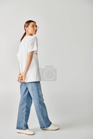 A young woman in a white shirt and jeans walks gracefully on a grey background.