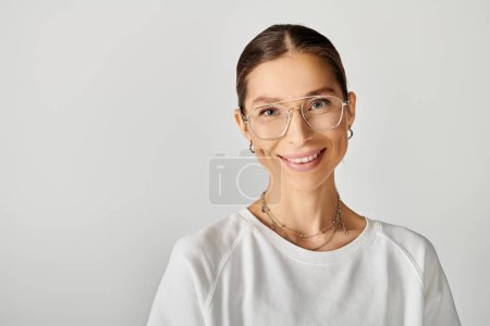 Une jeune femme en t-shirt blanc portant des lunettes sourit directement à la caméra sur un fond gris.