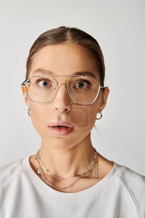 Une jeune femme en lunettes porte une expression surprise sur fond gris.