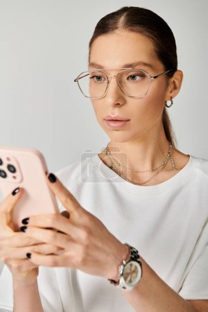 Eine junge Frau in weißem T-Shirt und Brille mit einem Handy vor grauem Hintergrund.