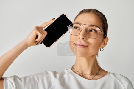 A woman in glasses holds up a cell phone with blank screen in studio