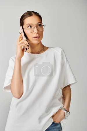 A stylish young woman in glasses chats on a cell phone against a grey backdrop, looking engaged and modern.
