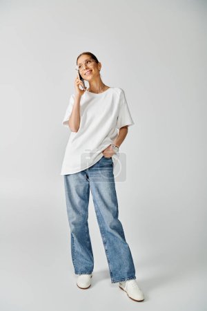 Téléchargez les photos : Une jeune femme vêtue d'une chemise blanche et d'un jean parlant sur un téléphone portable sur fond gris. - en image libre de droit