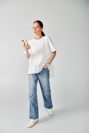 A stylish young woman in a white shirt and jeans casually texting on her cell phone against a grey backdrop.