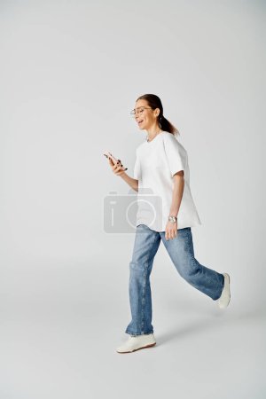 A young woman in a white t-shirt and glasses walks while focused on her cellphone screen.